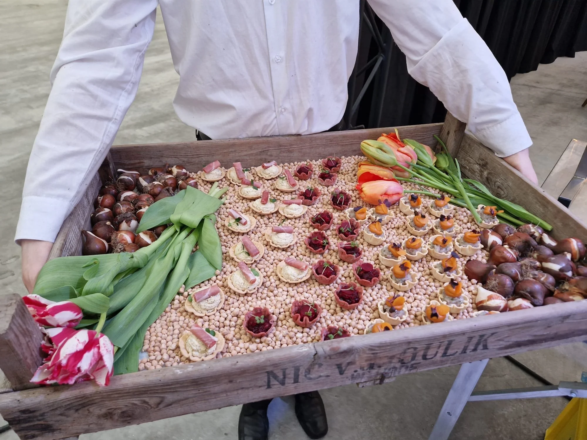 A wooden tray filled with floral arrangements, small round objects, tulips, and other decorative items.