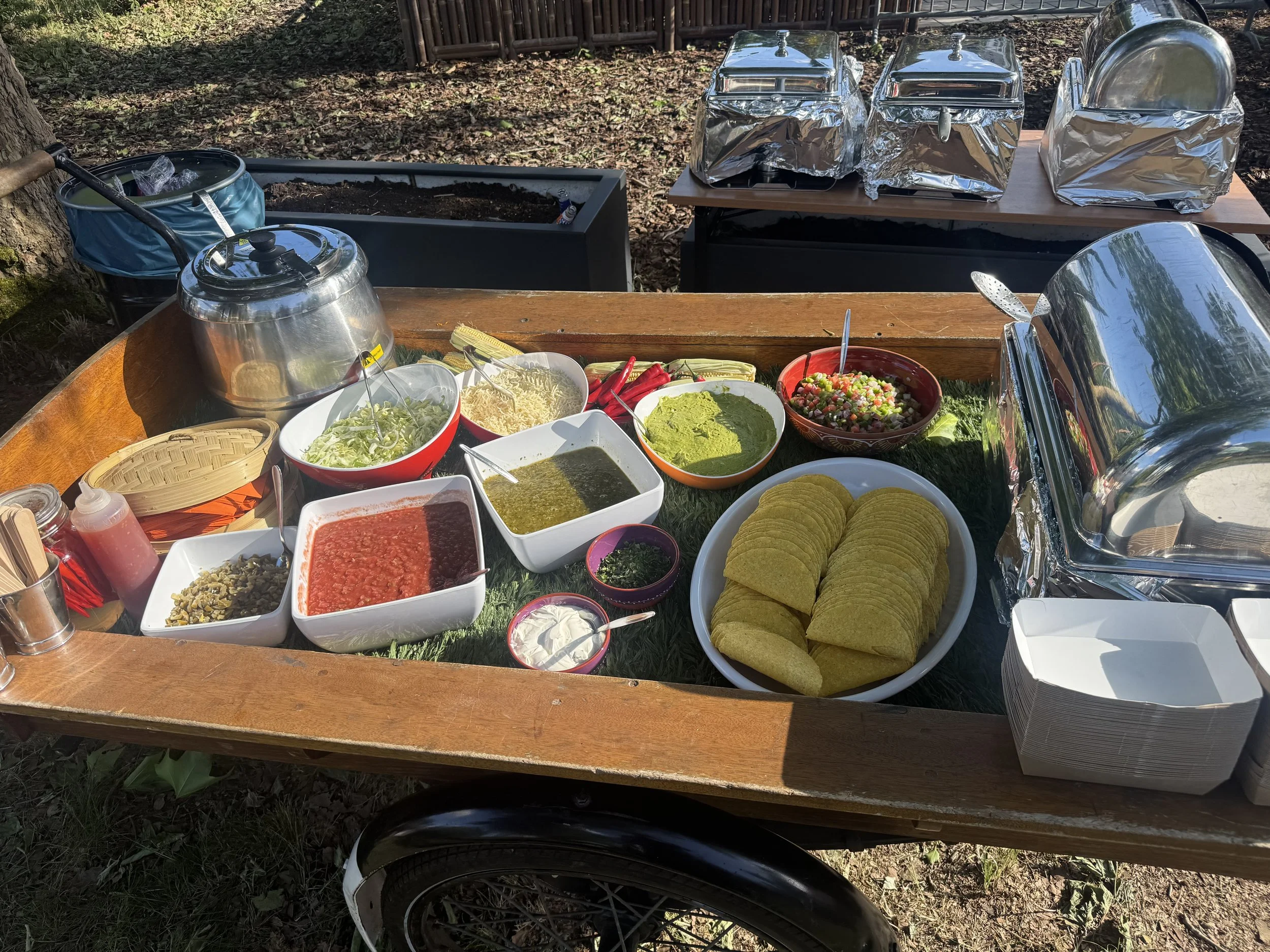 Outdoor taco bar setup with bowls of salsa, guacamole, shredded cheese, lettuce, tomatoes, and taco shells, along with warmers, napkins, and condiments.