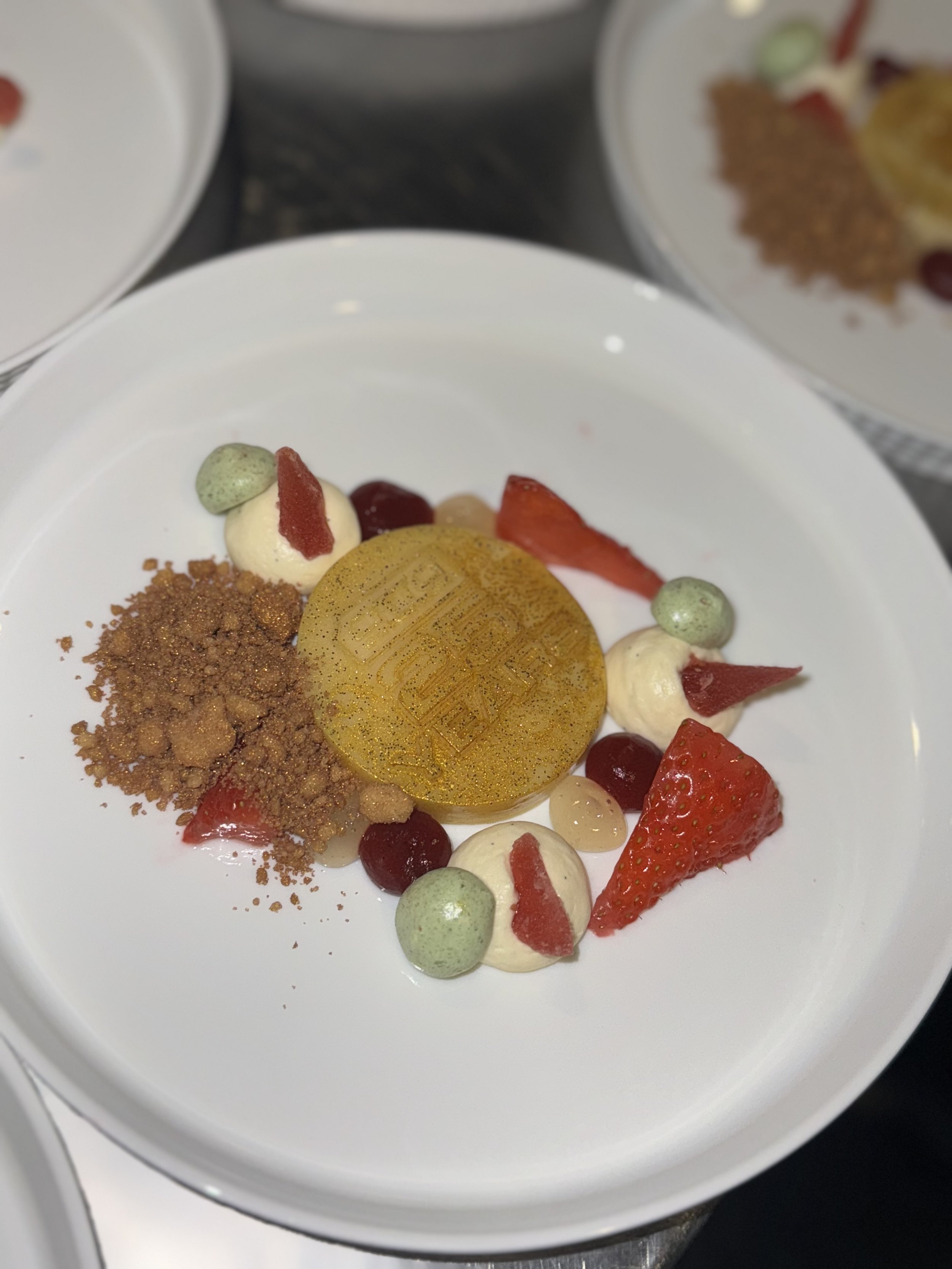 A plated dessert featuring a gold medallion with a geometric pattern, surrounded by strawberries, red and white sauces, small green and white dollops, and a crumbly brown topping.