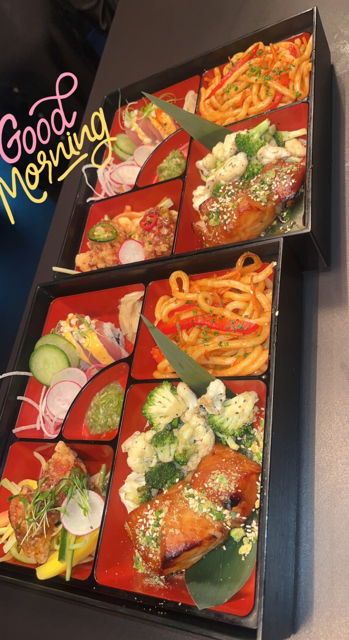 Two trays with multiple compartments of Japanese assorted dishes including sushi, noodles, and cooked vegetables, with a handwritten "Good Morning" greeting.