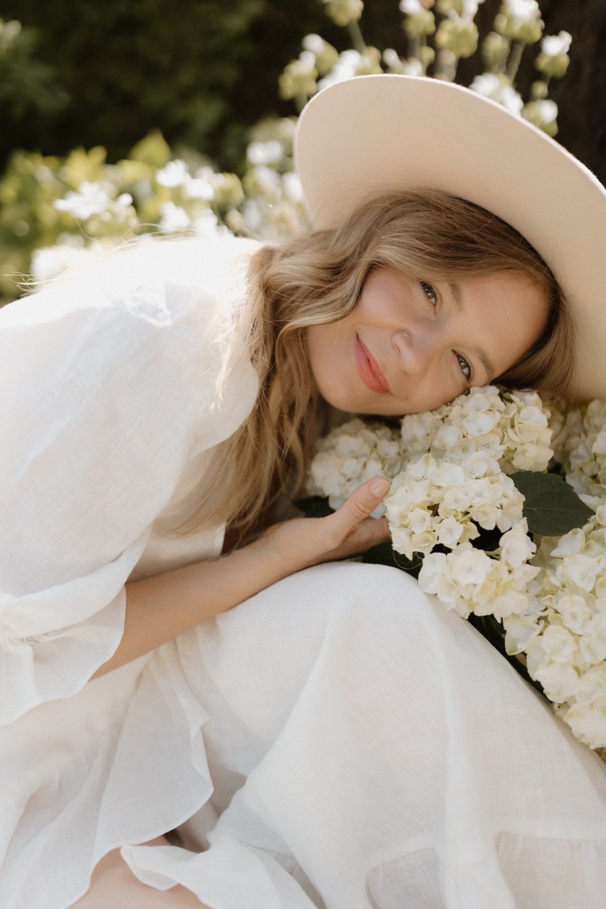 A woman with wavy blond hair lying down, smiling, resting her head on a large bouquet of white flowers, wearing a wide-brimmed hat and a white top.
