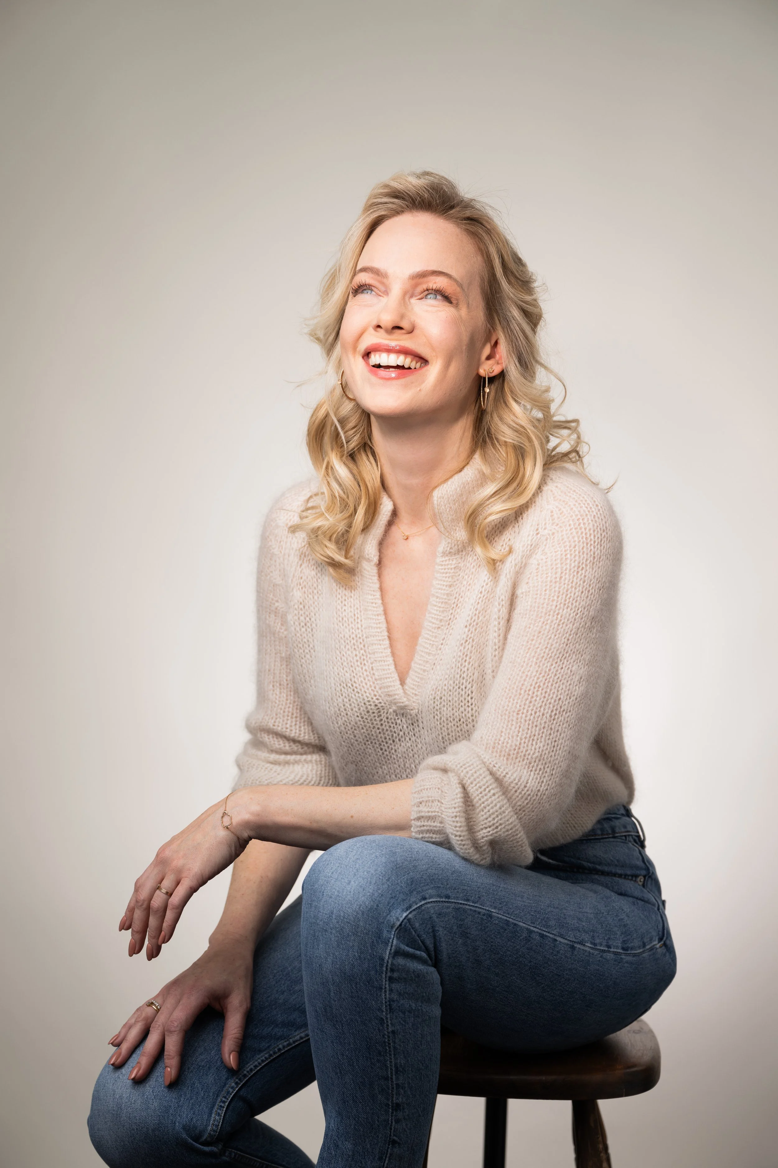 A smiling young woman with blonde, wavy hair wearing a beige knit sweater and hoop earrings, looking up and to the side.