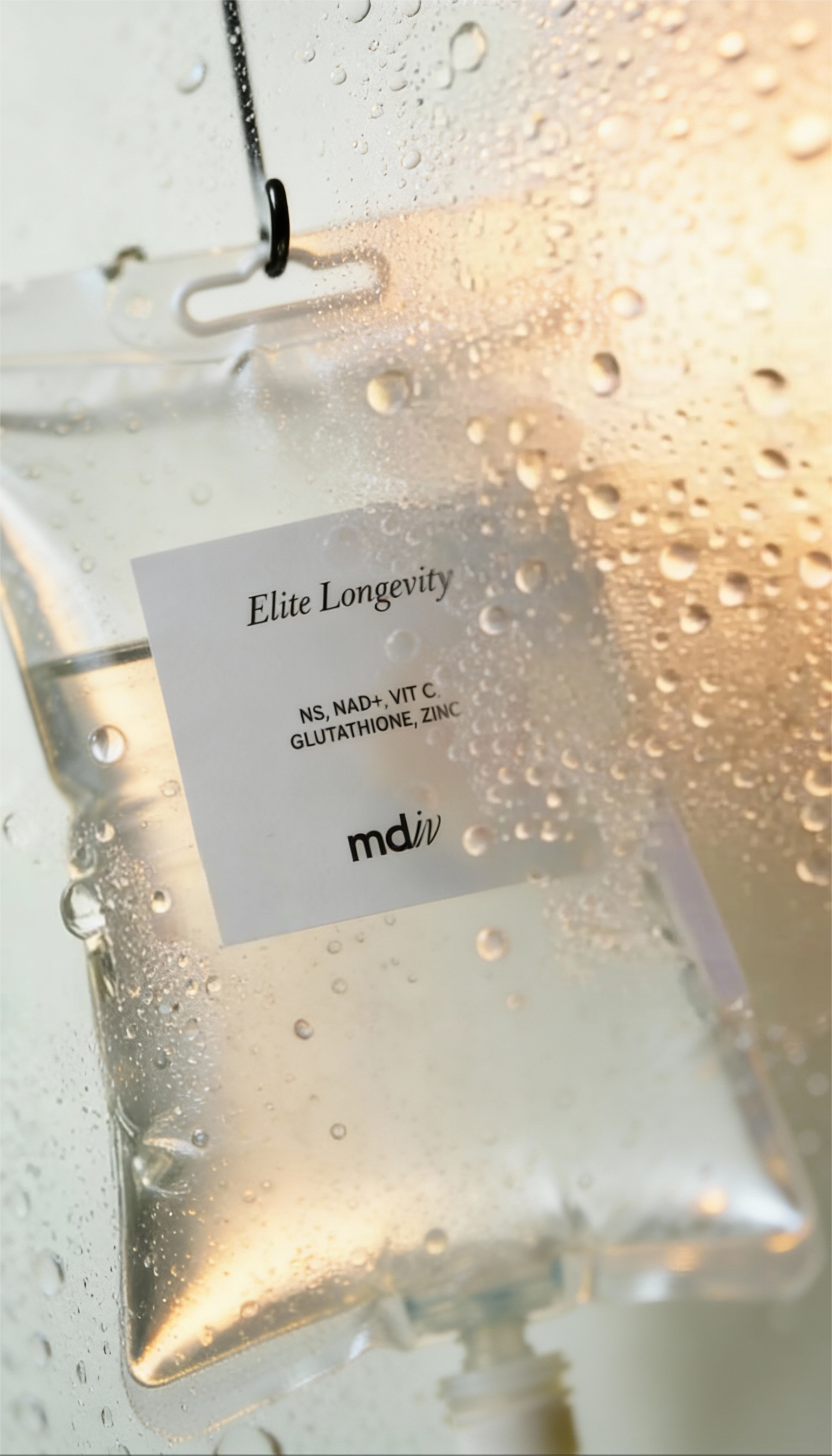 A clear IV bag with a label reading 'Elite Longevity' behind glass with condensation on the surface. The label mentions ingredients like NS, NAD+, VIT C, GLUTATHIONE, ZINC, and has the logo 'md iv'.