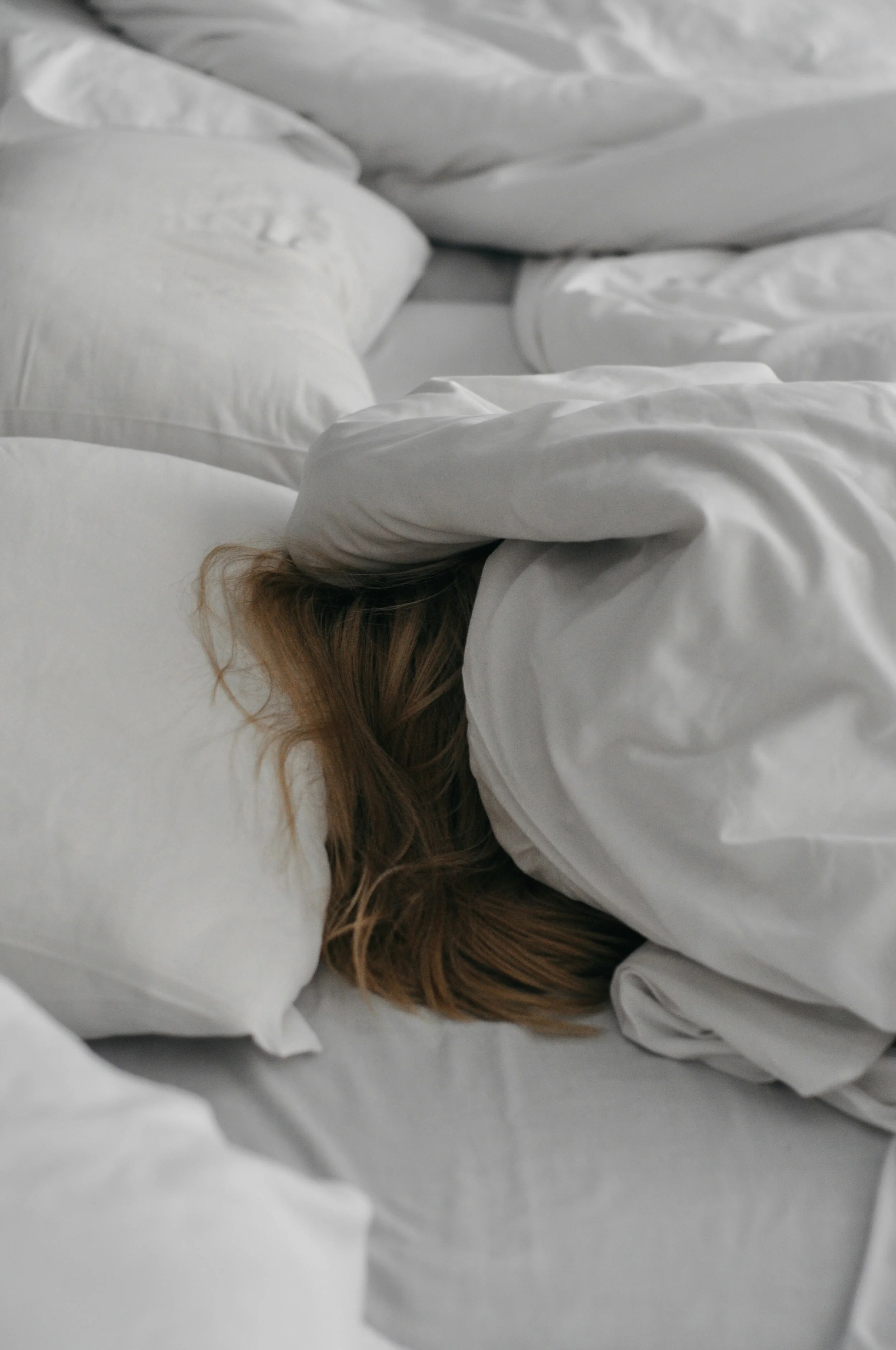 A person with brown hair sleeping on a white pillow in a bed with white sheets and blankets.