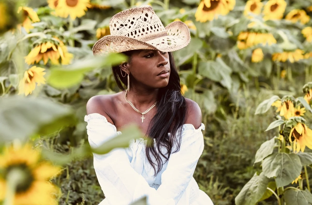 A woman with long black hair wearing a straw hat, white off-the-shoulder dress, silver necklace with a cross, and hoop earrings, sitting in a sunflower field.