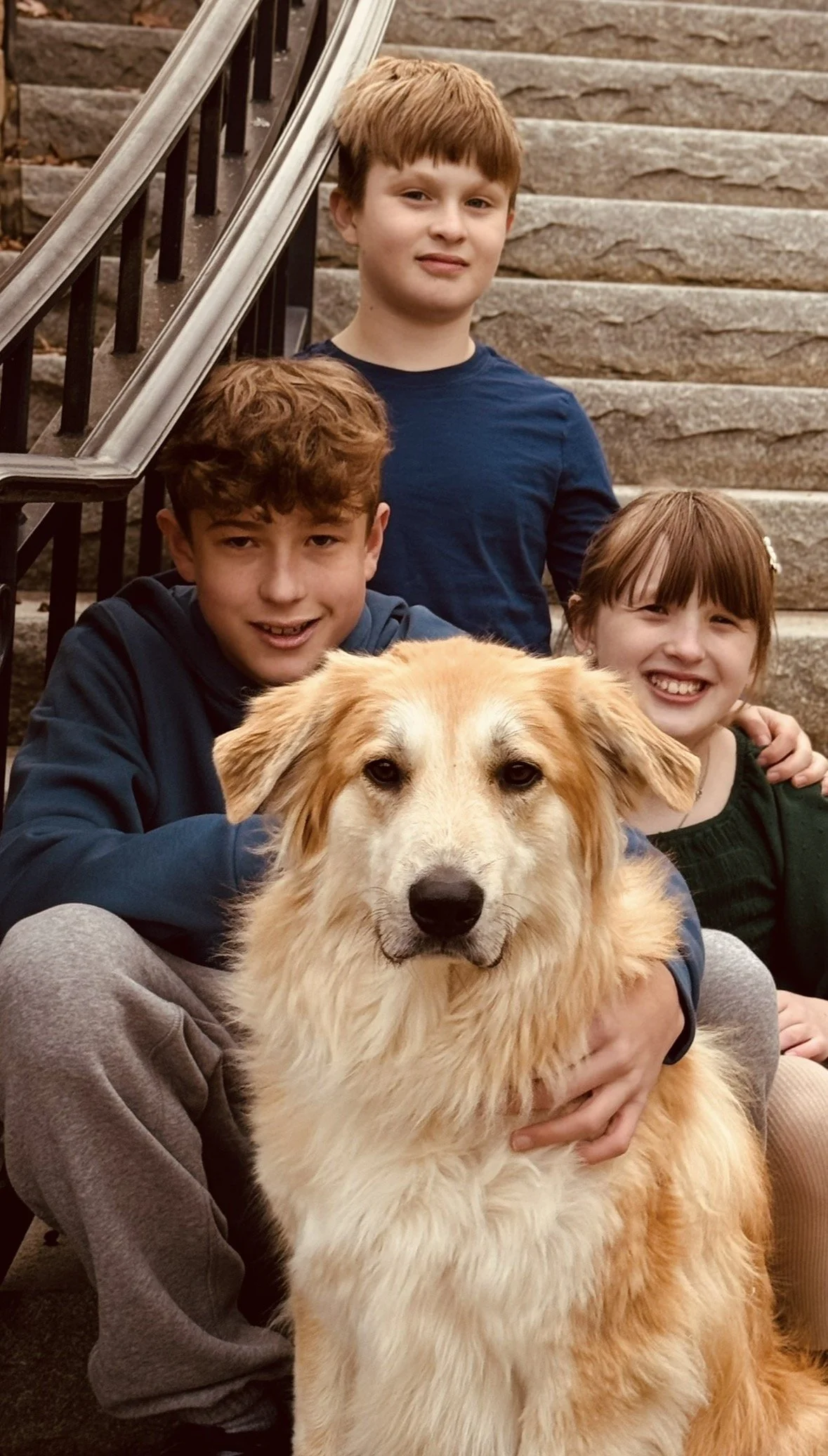 Three children and a golden retriever dog sitting on stone steps outdoors. The kids are smiling, with the dog in front.