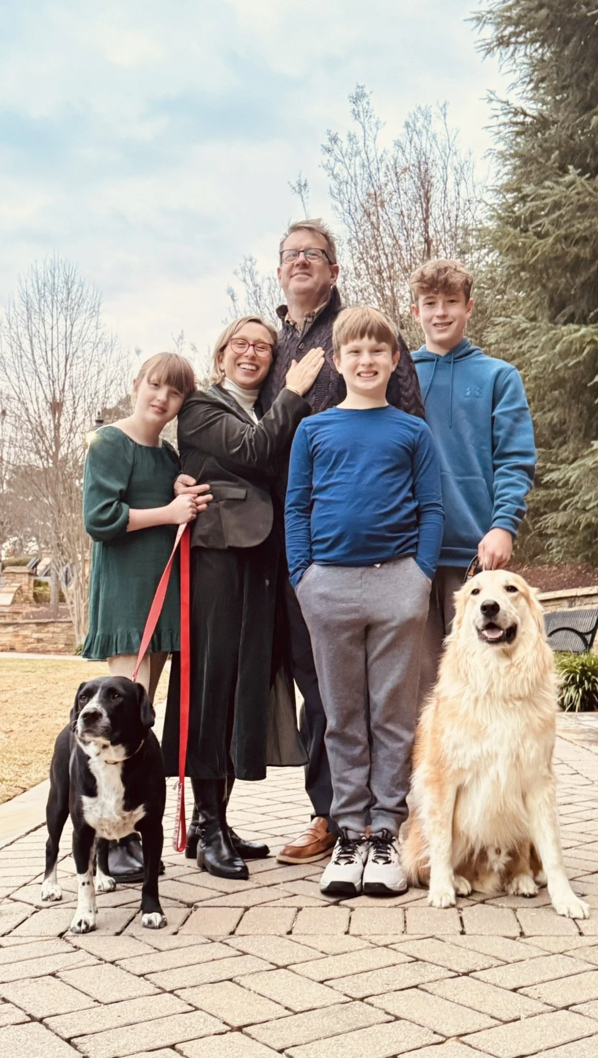 A family of five with two dogs, two adults, and three children posing outside on a brick pathway in a backyard with trees and a stone wall in the background