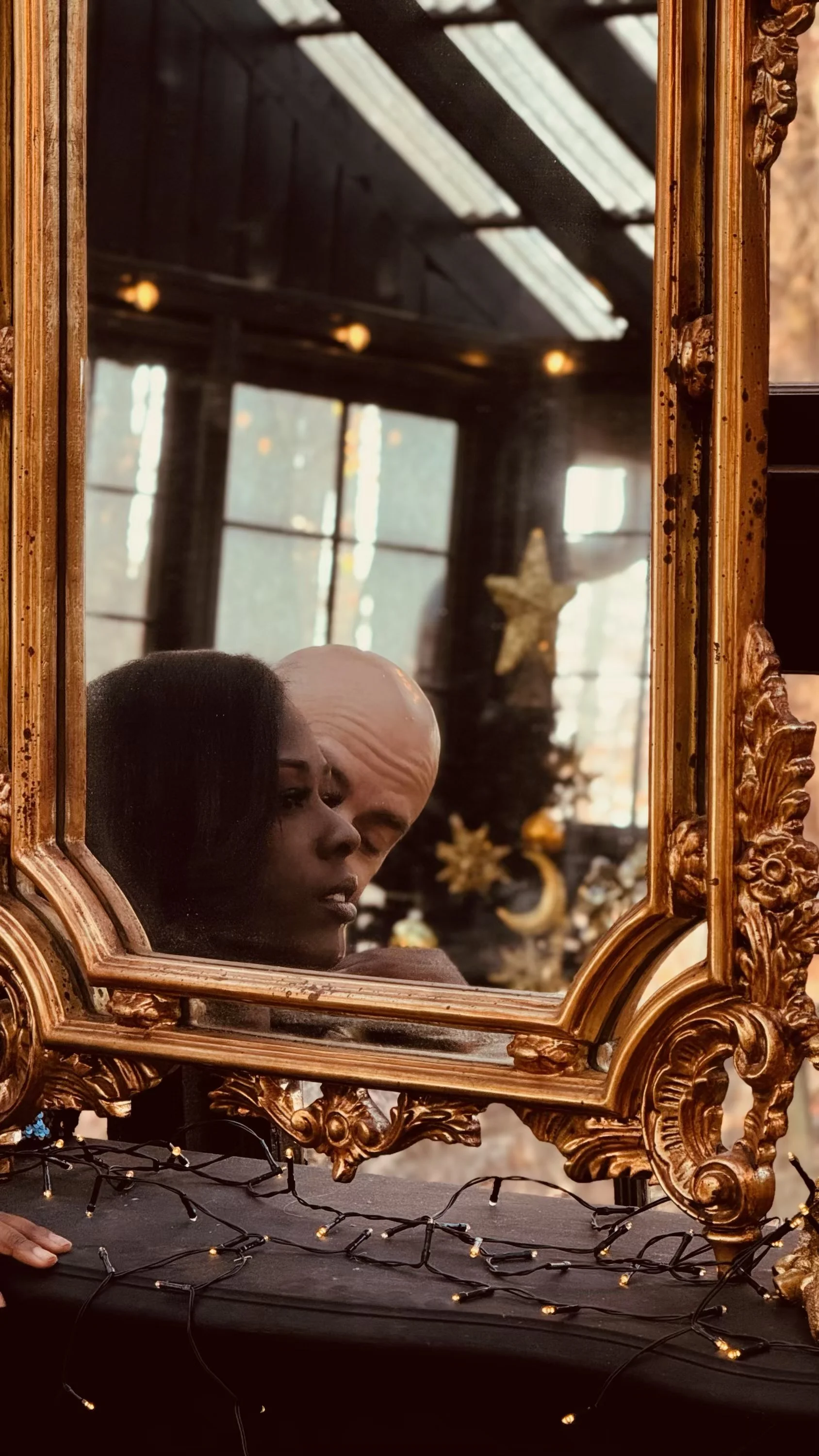 A reflection of a woman with dark hair and a man with a bald head in an ornate gold mirror, with a decorated Christmas tree behind them.