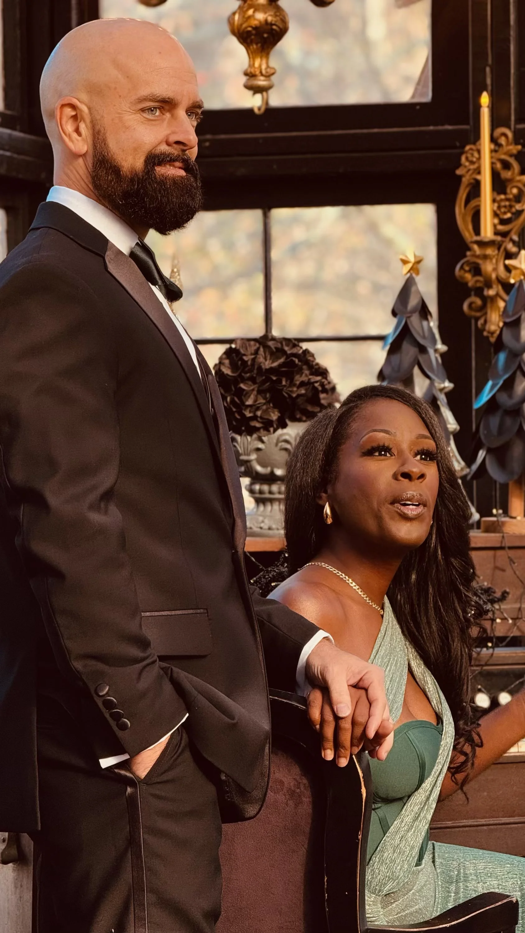 A man in a tuxedo standing behind a seated woman in a green dress, holding her hand, in an ornate, warmly lit indoor setting with decorative objects and candles.