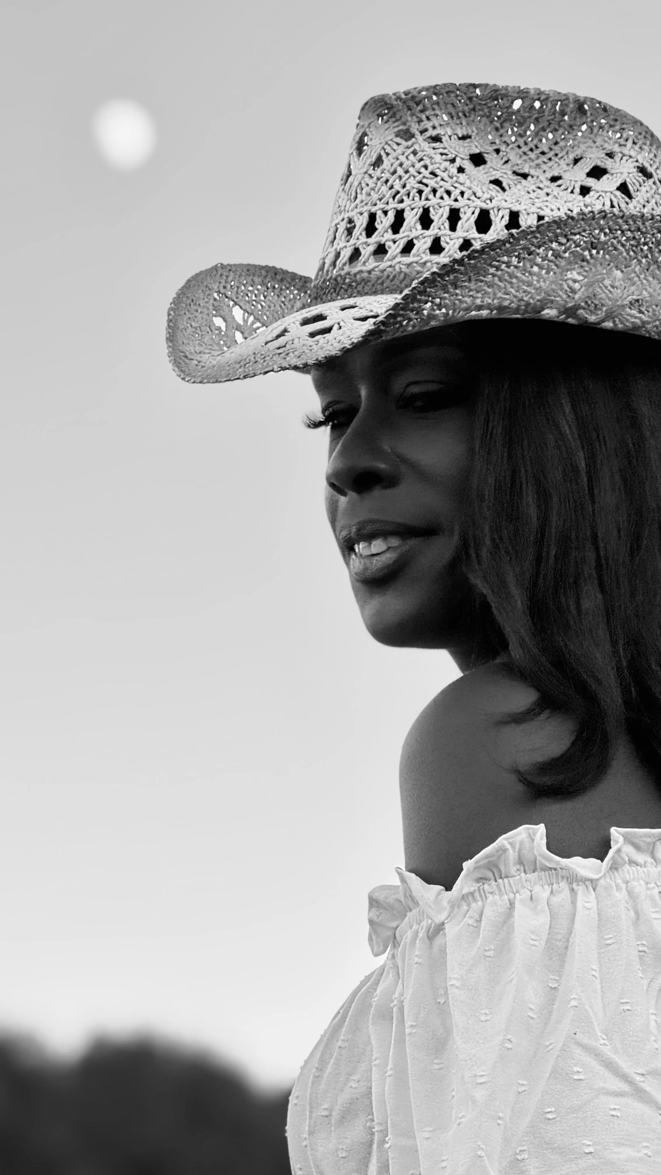 A woman wearing a wide-brimmed woven hat and an off-the-shoulder white top, smiling softly with a blurred natural background.