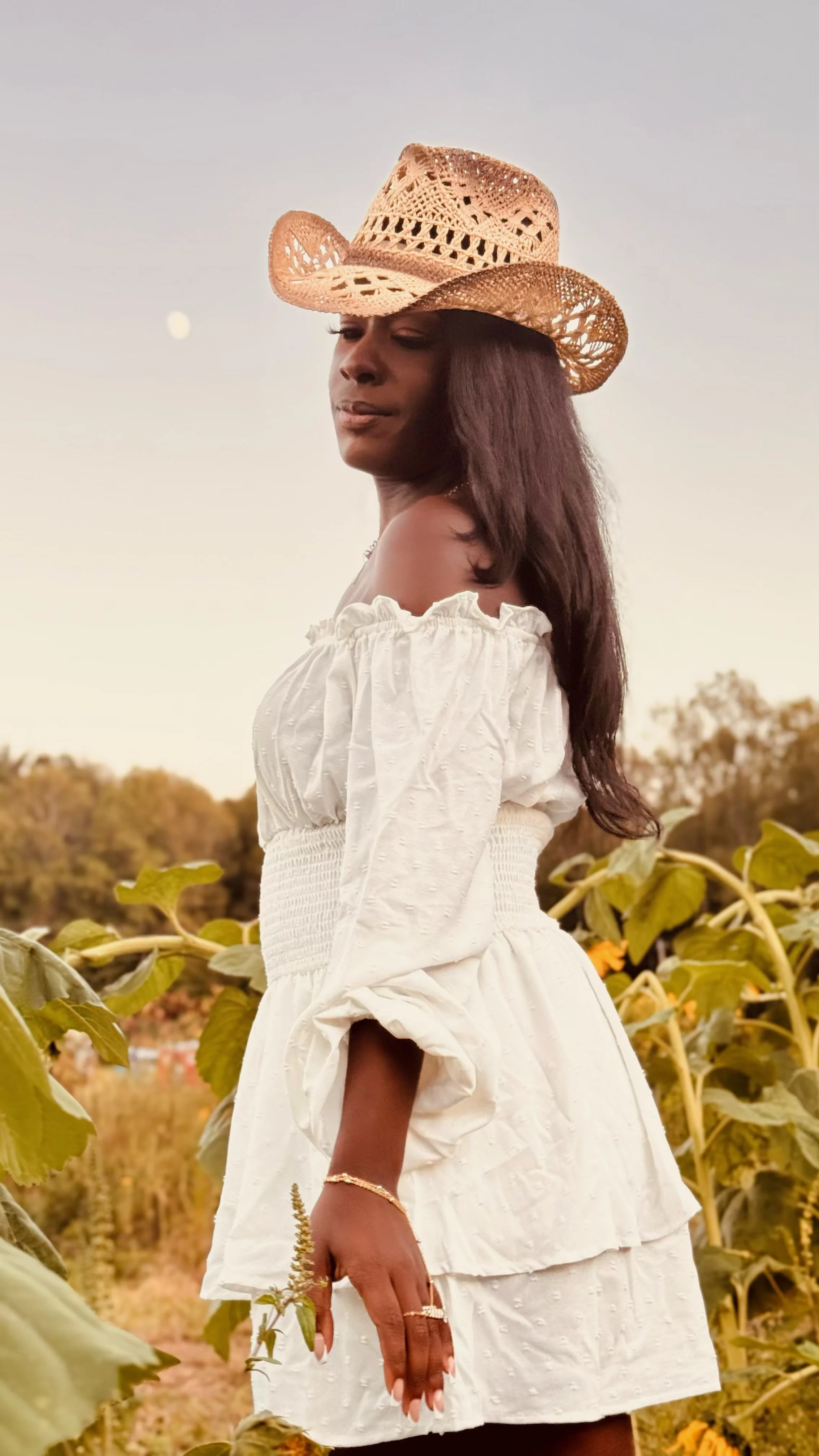 A woman in a white off-the-shoulder dress and wearing a straw cowboy hat standing outdoors among green foliage with a clear sky and the moon visible in the background.