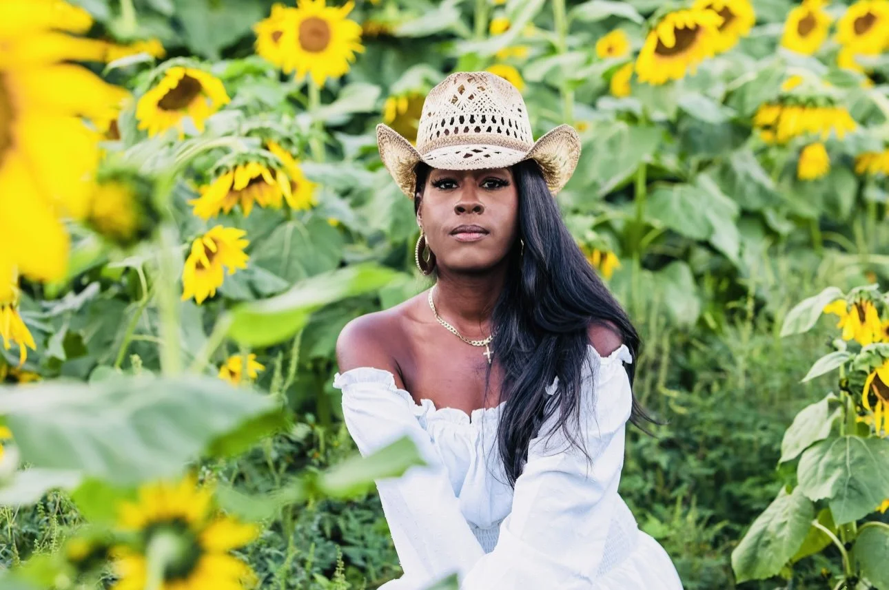 A woman with long black hair, wearing a straw hat and an off-the-shoulder white dress, sitting among sunflowers in a garden.