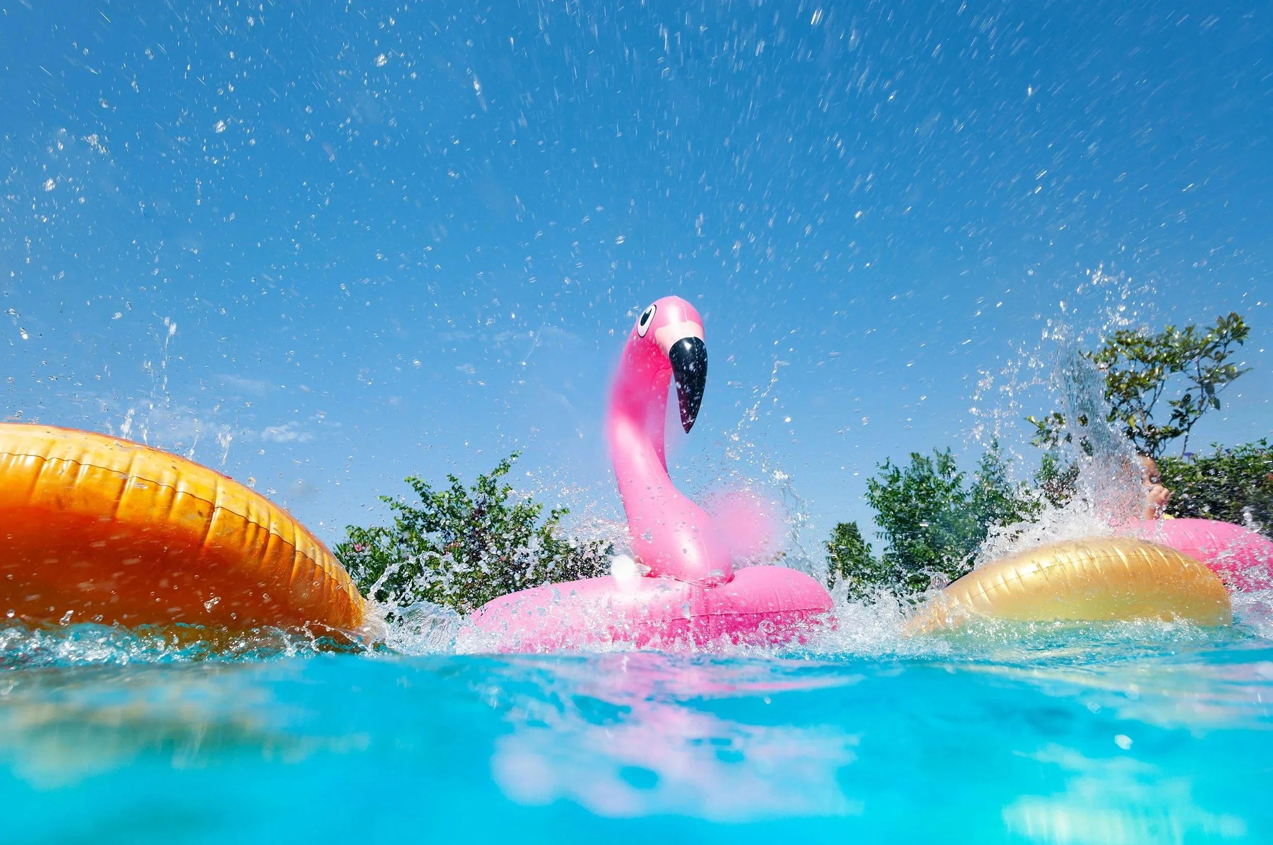 Pink inflatable flamingo float in a pool with splashing water, under a clear blue sky with trees in the background.