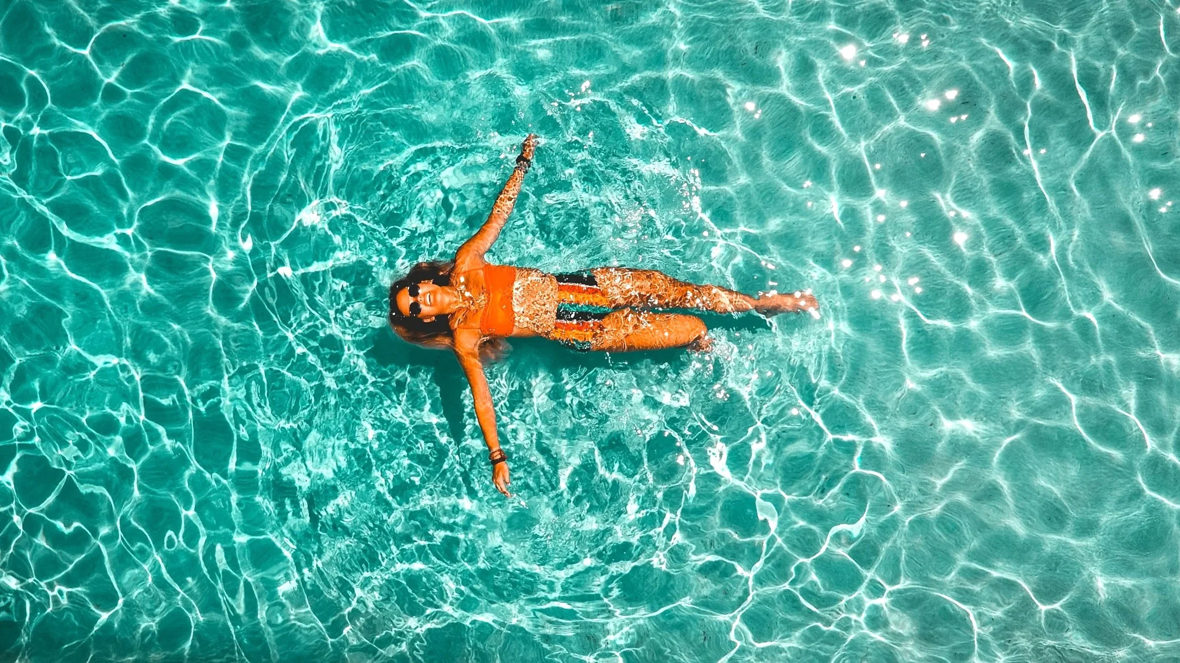 A woman in a bright orange swimsuit and sunglasses is floating on her back in a clear, turquoise swimming pool, relaxing and enjoying the water.