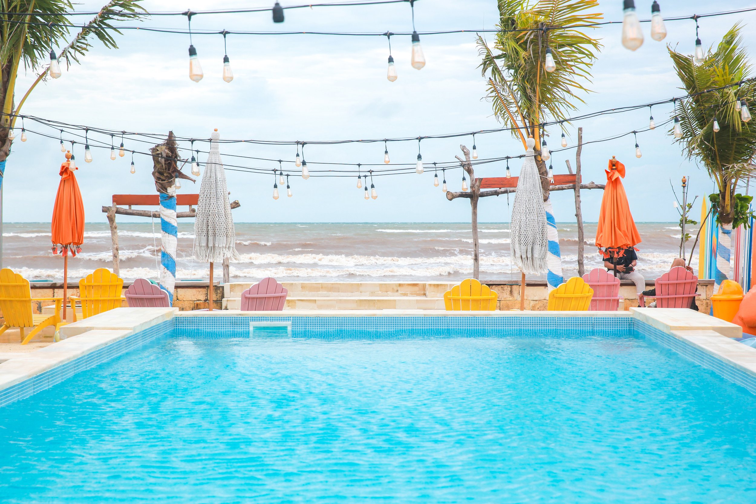 Swimming pool with a view of the beach and ocean, colorful Adirondack chairs, umbrellas, and string lights, with palm trees.