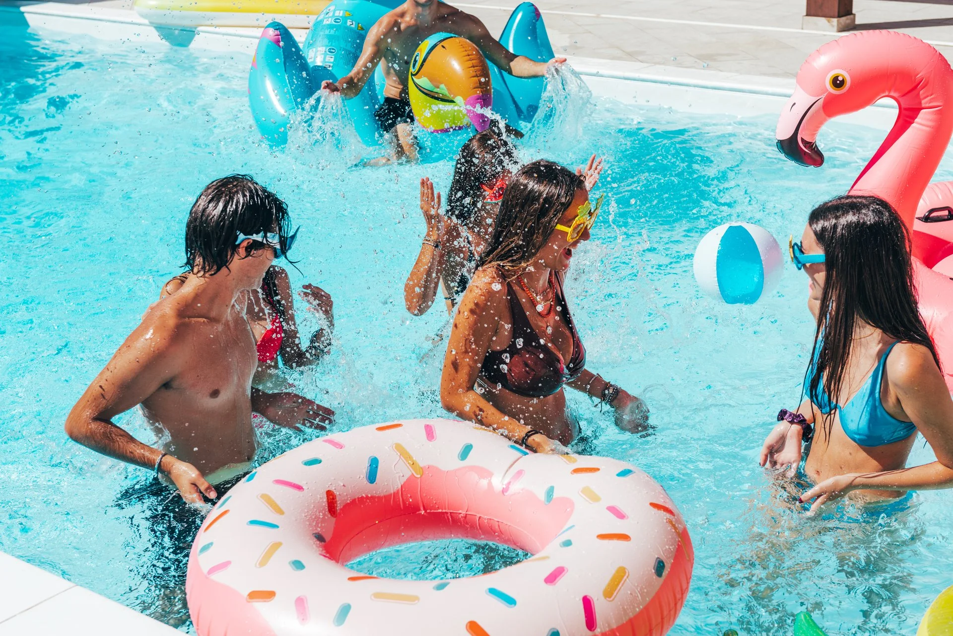 Children playing in a swimming pool with floatation toys, including a donut-shaped float with sprinkles and a pink flamingo float, enjoying a sunny day.