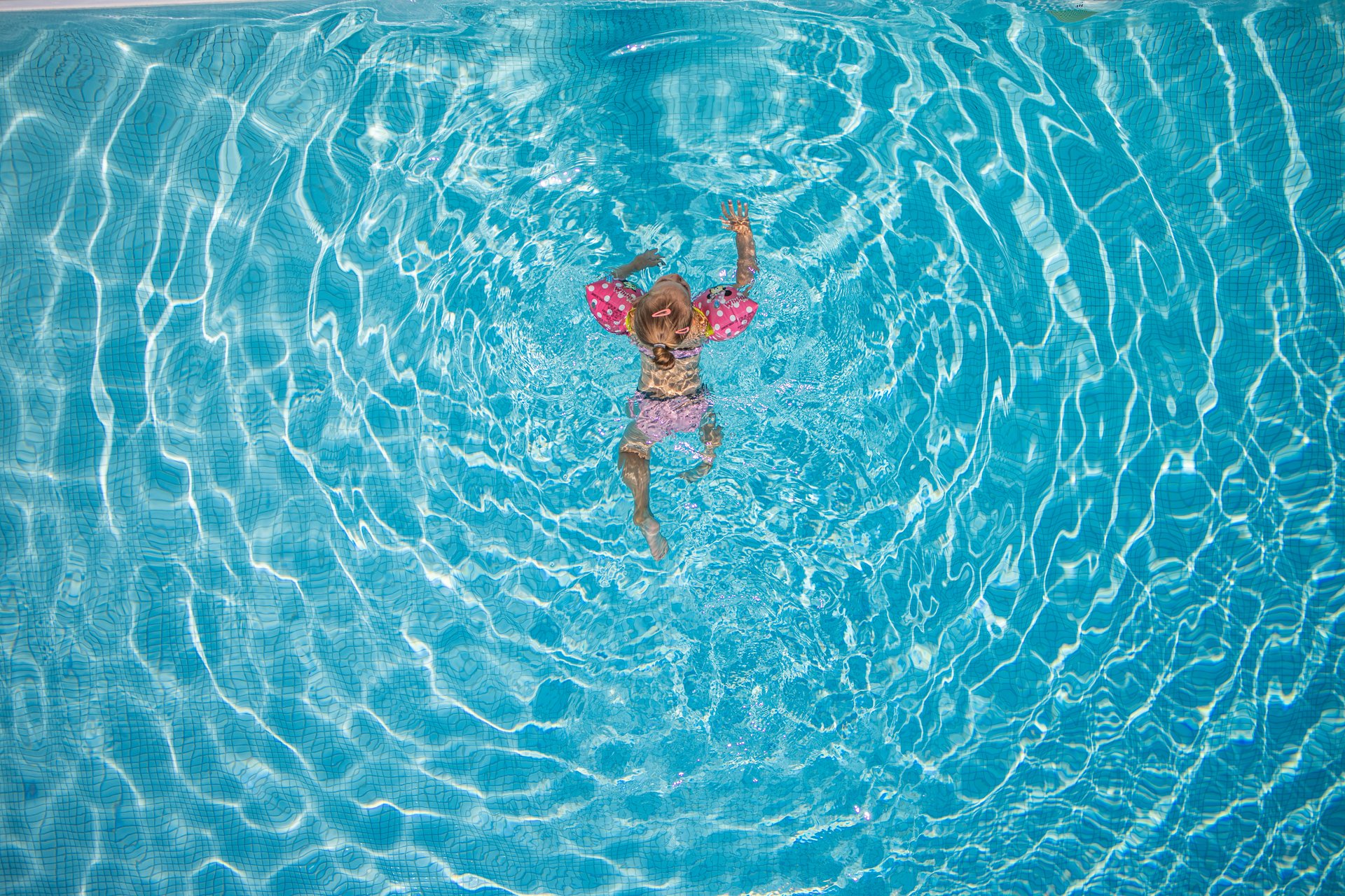 A young girl in a pink swimsuit with polka dots swimming in a clear blue pool, viewed from above.