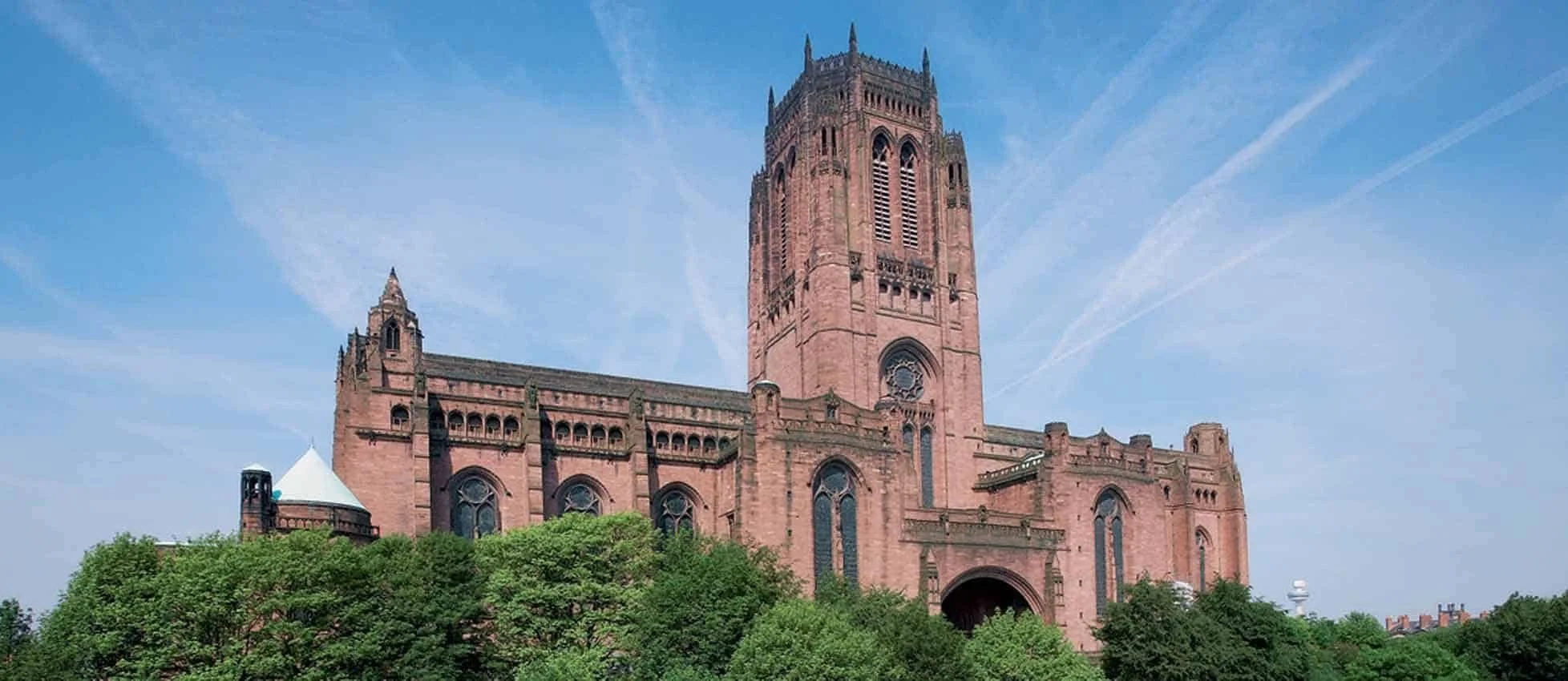 A large historic stone church with a tall tower, red or pinkish stone construction, gothic architectural features, and surrounded by green trees under a blue sky with contrail streaks.