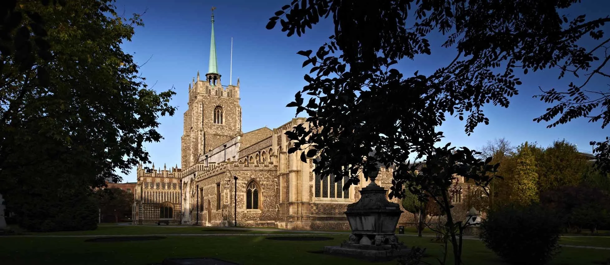 A historic stone church with a tall steeple, surrounded by green trees and a manicured lawn under a clear blue sky.