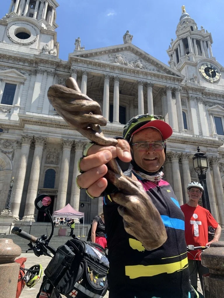 Man in cycling gear in front of cathedral, holding a bronze sculpture in the shape of two hands holding a baton
