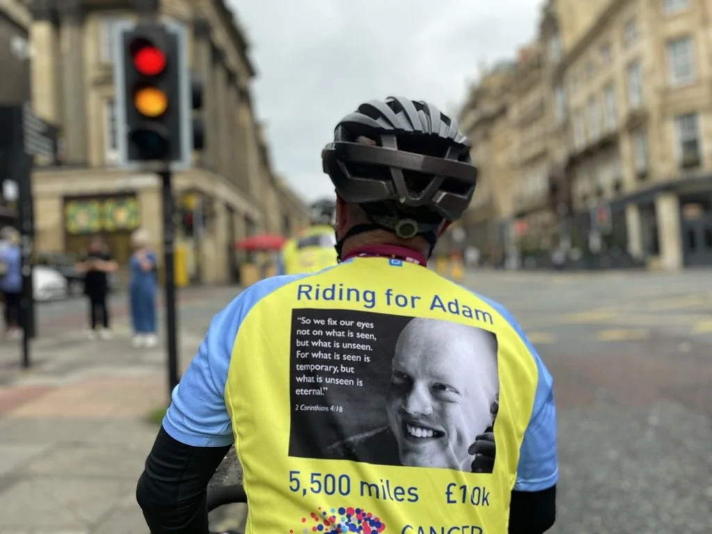 A cyclist with a helmet wearing a yellow and blue jersey that reads 'Riding for Adam' on a city street. The jersey displays a quote from 2 Corinthians 4:18, a distance of 5,500 miles, a donation amount of £10,000, and the word 'CANCER.' The background shows a red traffic light, a few people, and city buildings.
