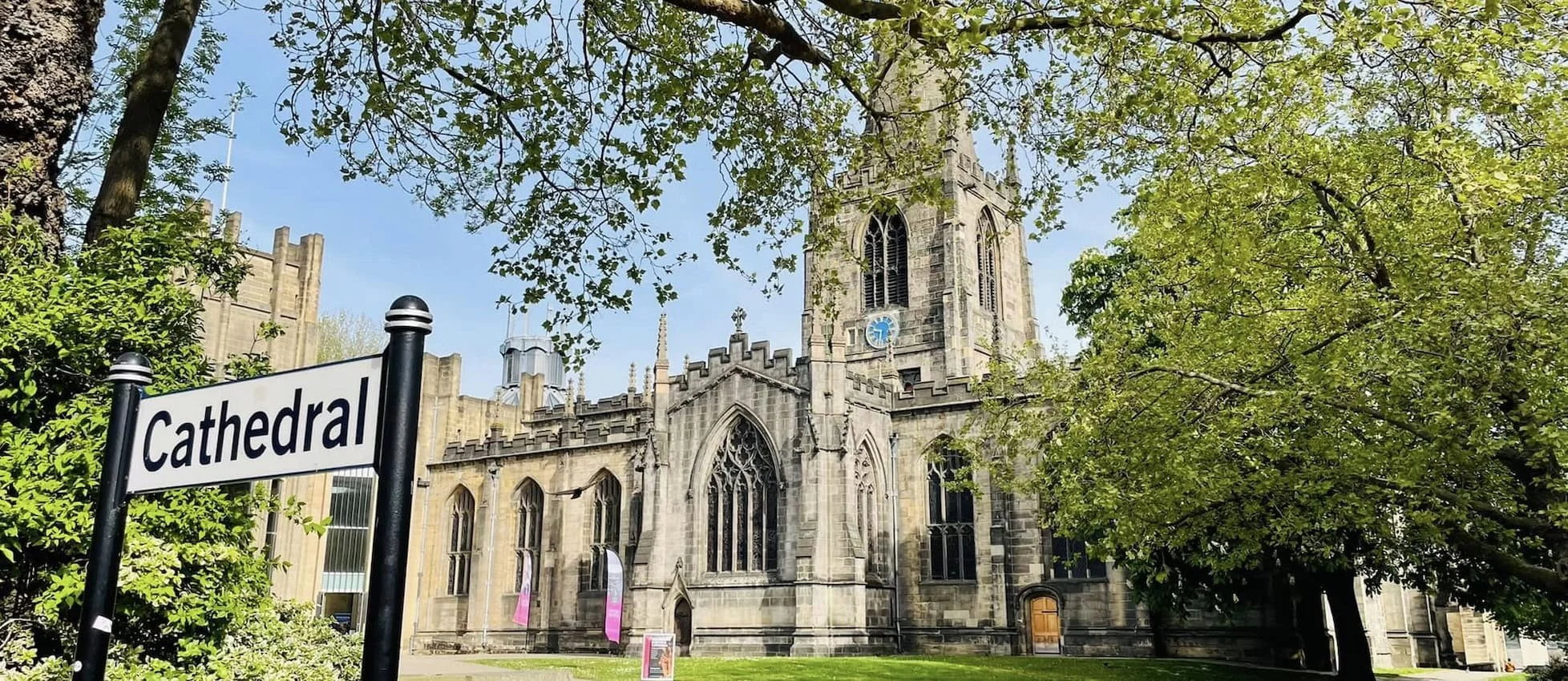 View of a historic stone cathedral with a tall tower, arched windows, surrounded by green trees, and a sign that reads 'Cathedral' in the foreground.