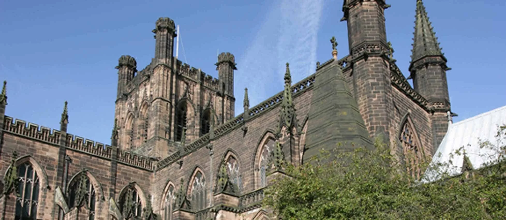 The image shows a large Gothic-style church with tall spires and brick walls against a blue sky. Green trees partially cover the lower part of the building.