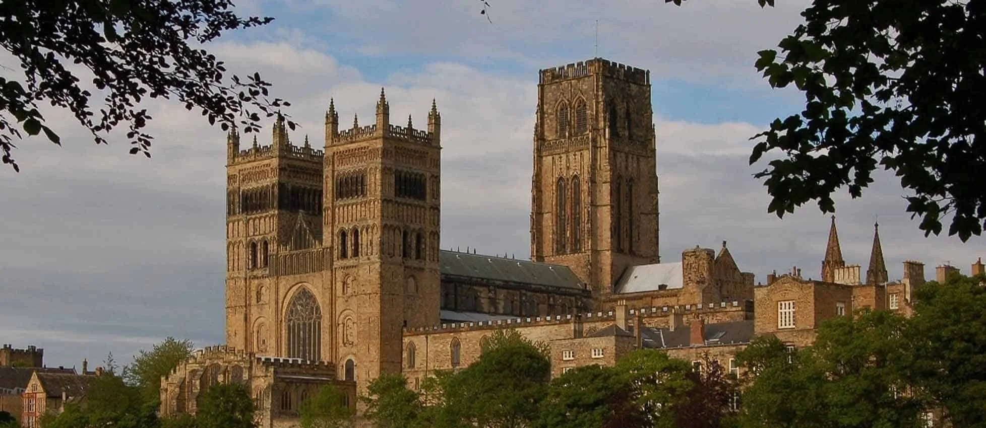 Sunlit view of a historic stone cathedral with two towers, surrounded by trees, under a partly cloudy sky.