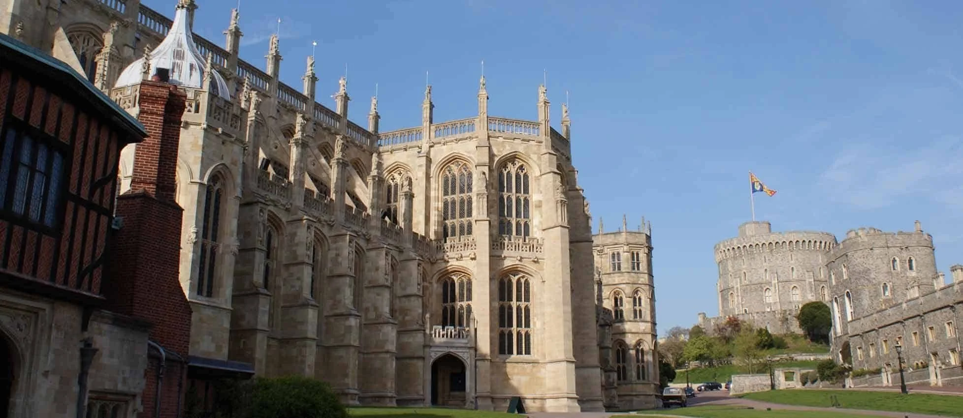 View of Windsor Castle with a blend of medieval and Gothic architecture, stone walls, and a flag on top, under a clear blue sky.