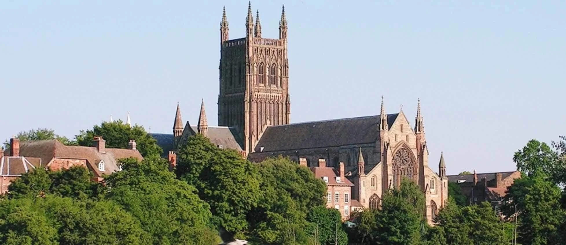 A large Gothic-style church with a tall central tower, surrounded by green trees and smaller buildings, under a clear blue sky.