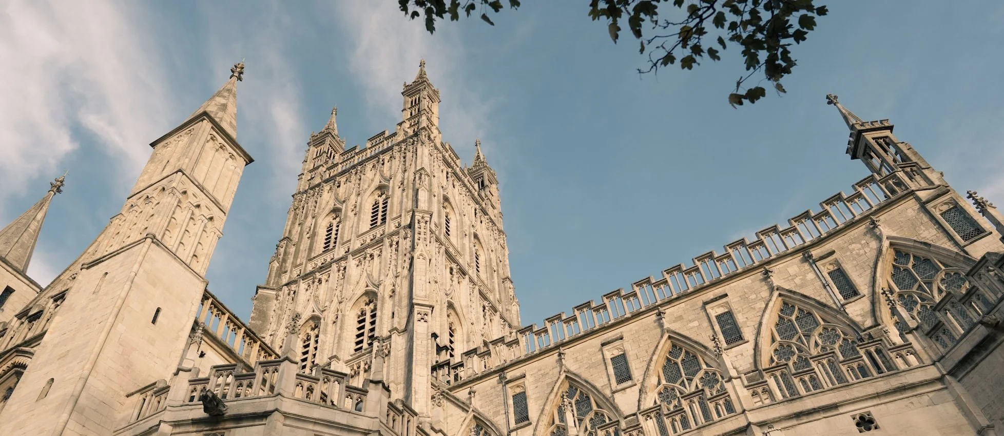 Low-angle view of a Gothic cathedral with tall, ornate spires and large stained glass windows, against a blue sky with some clouds and tree branches at the top.