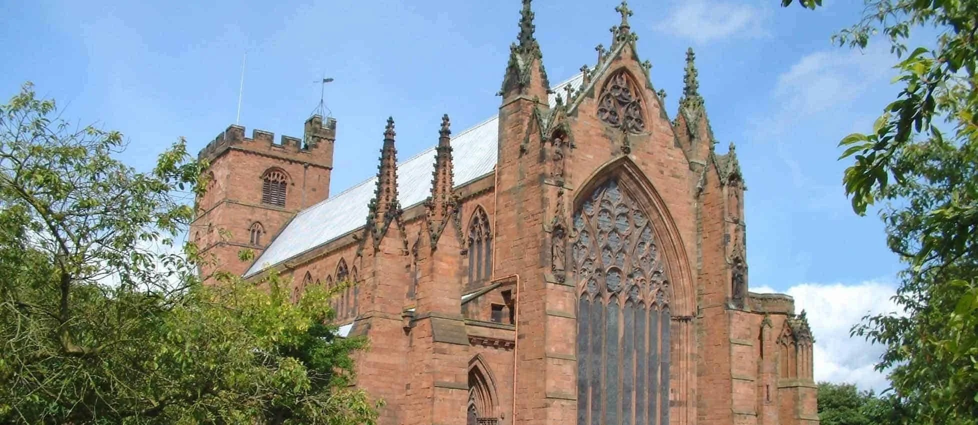 A large Gothic-style church made of red bricks with stained glass windows and pointed arches, surrounded by green trees under a blue sky.