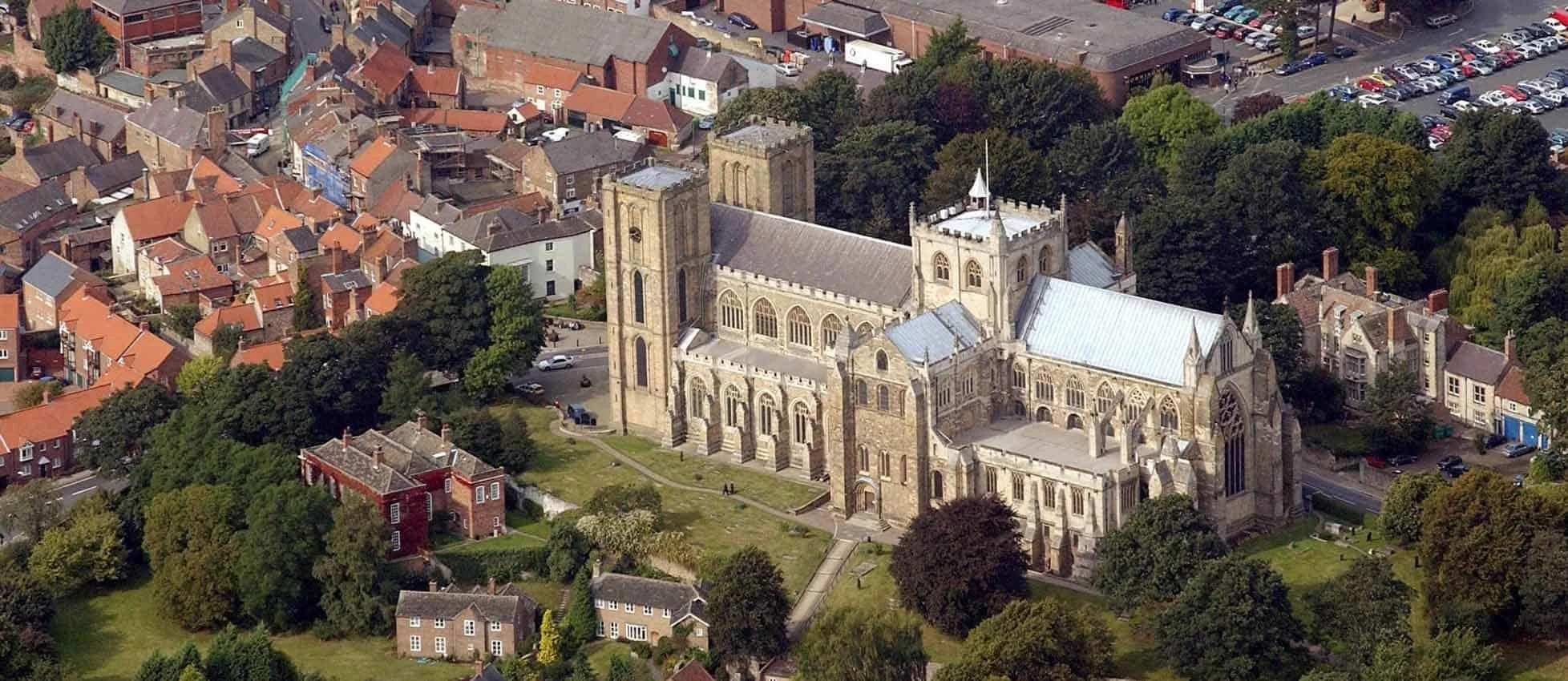 An aerial view of a historic stone church with two tall towers, surrounded by residential houses and trees in a small town.