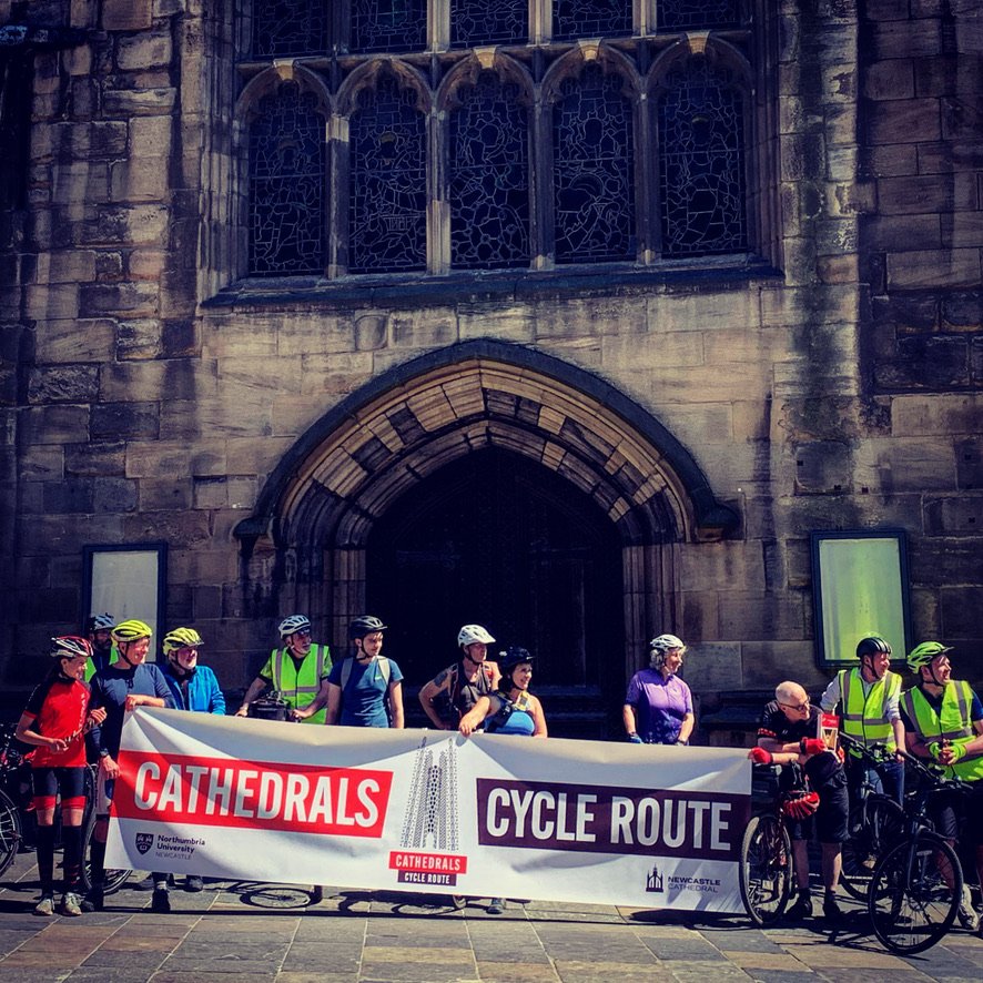 A group of cyclists holding a banner that reads 'Cathedrals Cycle Route' standing in front of a large, historic cathedral with stained glass windows and an arched entrance.