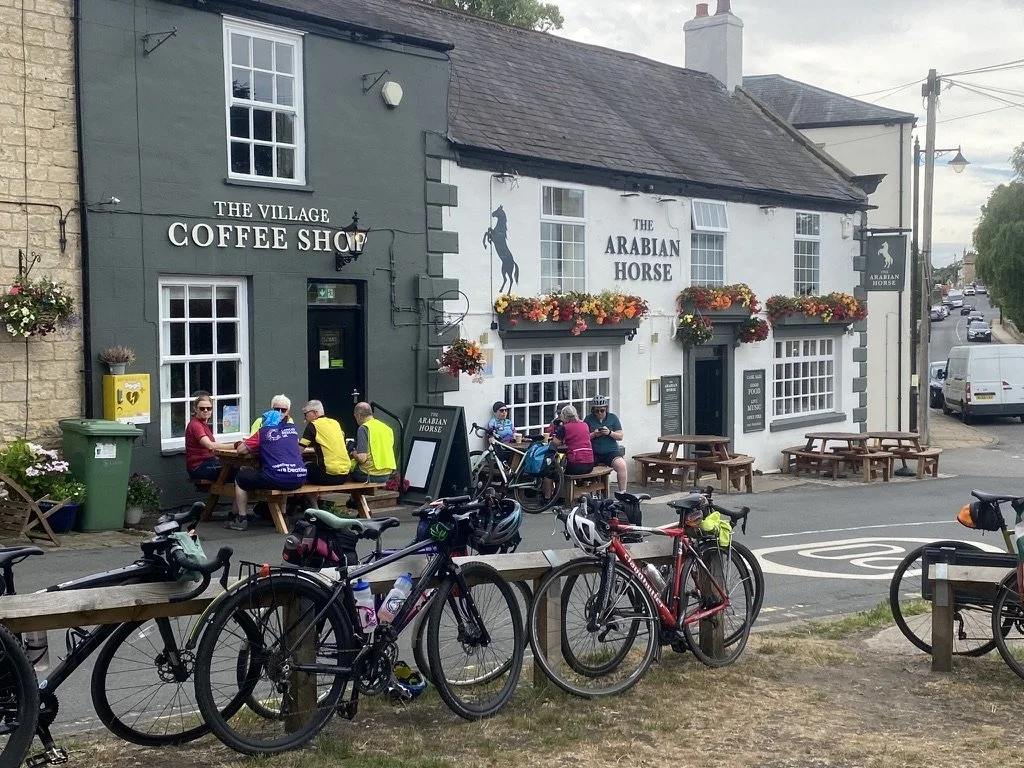A quaint corner in a village with a coffee shop called "The Village Coffee Shop" and a pub called "The Arabian Horse." People are seated outside at wooden tables, some with bicycles parked nearby. The storefronts are decorated with flower boxes, and the sky is partly cloudy.