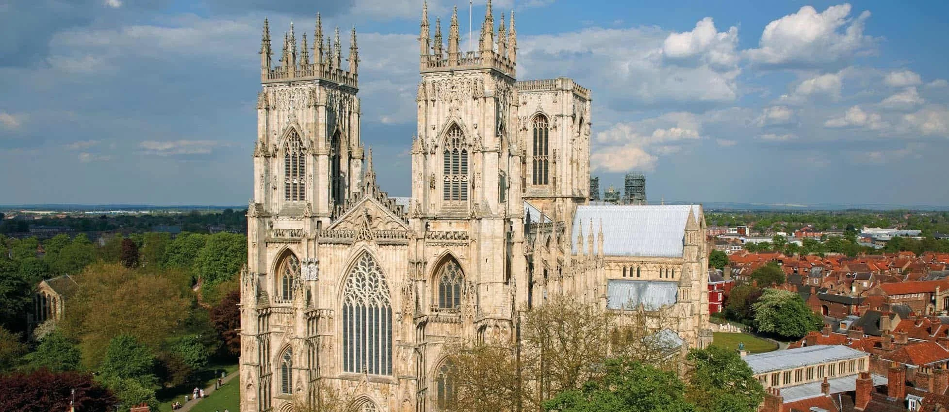A historic Gothic-style cathedral with two tall spires, large stained glass windows, and intricate stone carvings, set against a partly cloudy sky with green trees and a town with red-roofed buildings in the background.