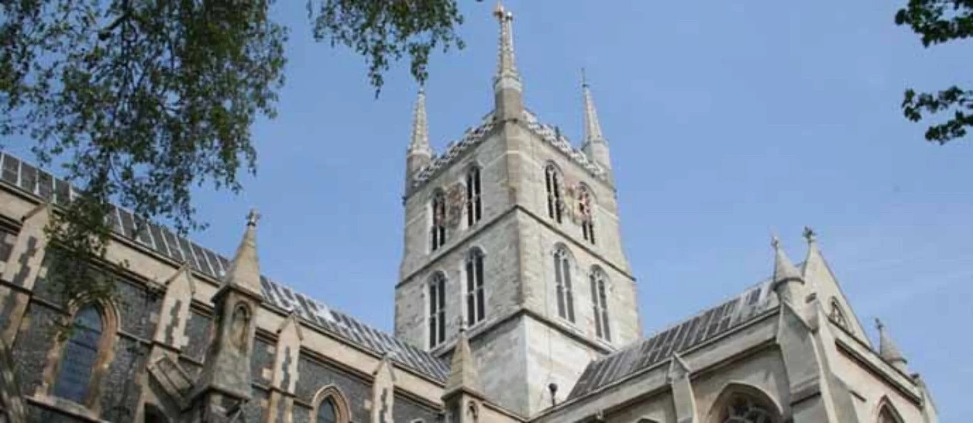 A tall, historic stone church with pointed spires and large arched windows, set against a clear blue sky with leafy tree branches partially framing the image.