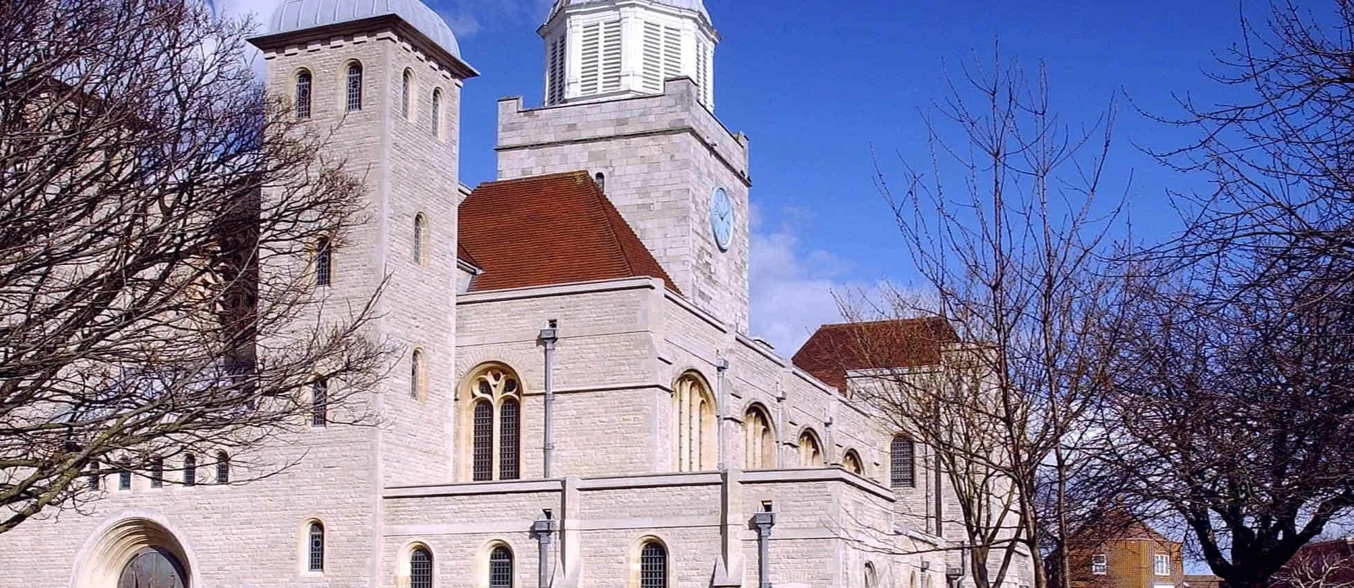 A historic stone church with a clock tower, red-tiled roofing, and leafless trees in the foreground, set against a bright blue sky.