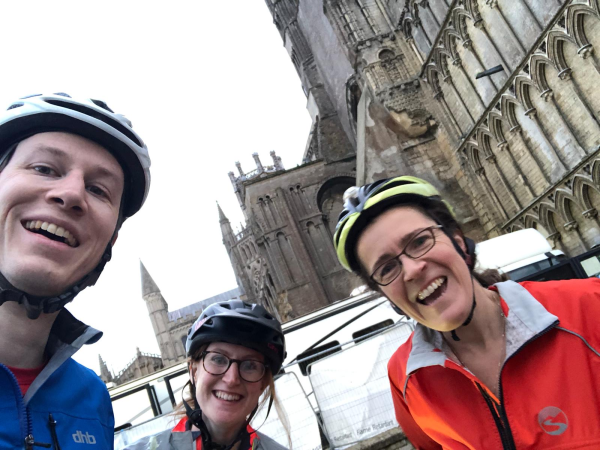 A blue directional sign for the National Cycle Network Centre is attached to a black pole in front of a historic stone church or cathedral with large stained glass windows and gothic architectural features.