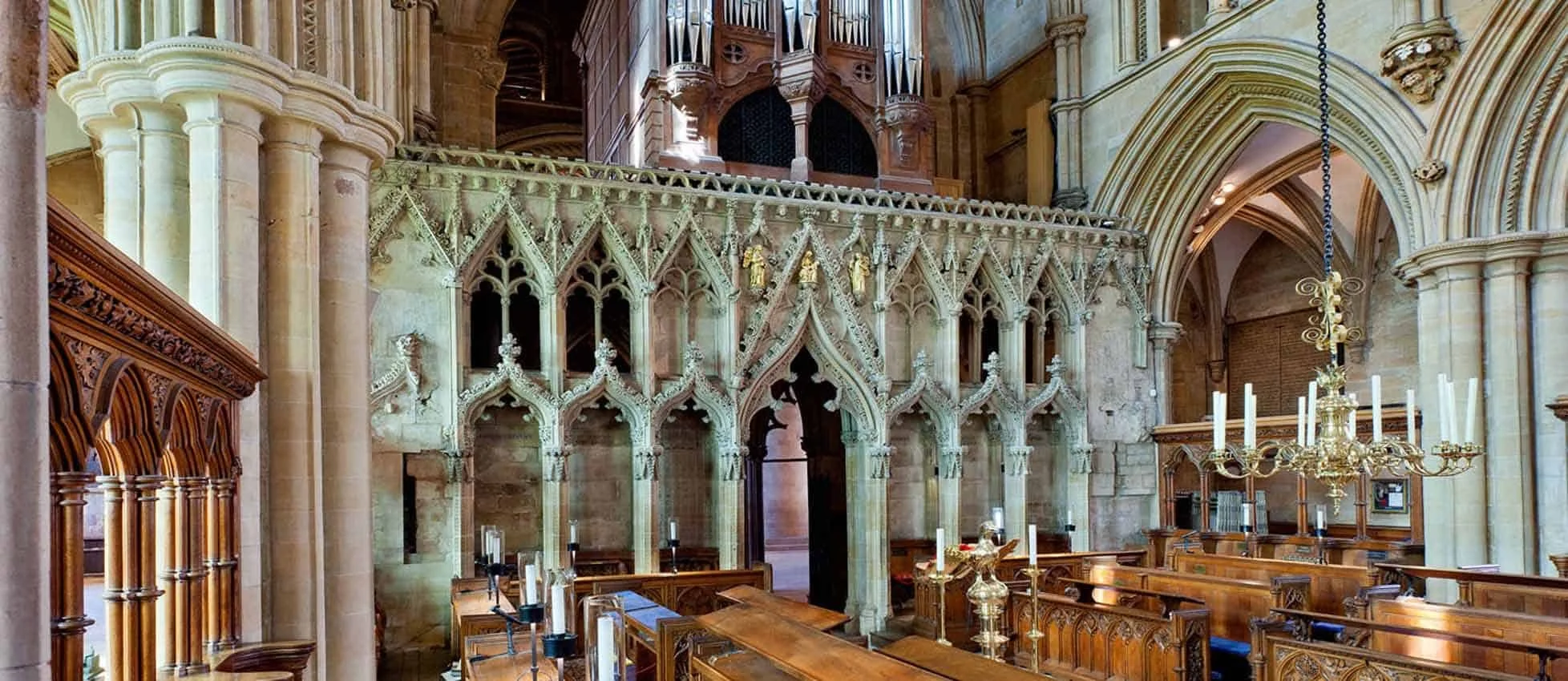 Interior of a historic church or cathedral featuring detailed Gothic architecture with intricate stone carvings, wooden pews, and a large chandelier hanging from the ceiling.