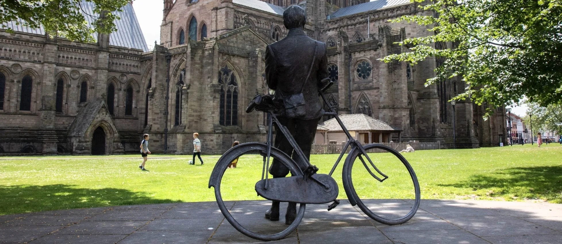Bronze statue of a man with a briefcase walking with a bicycle in front of a large Gothic-style church, with children playing in the background and trees shading the scene.