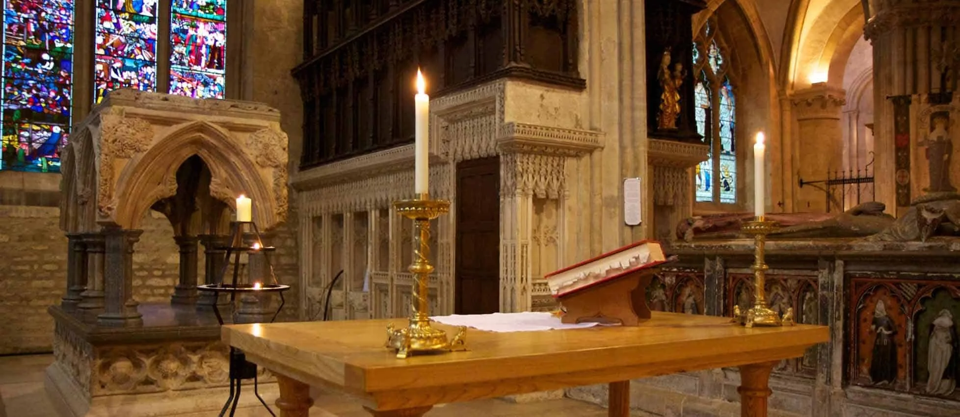 Inside a historic church or cathedral, featuring stained glass windows, an altar with lit candles, a large open Bible, and intricate stone and woodwork.