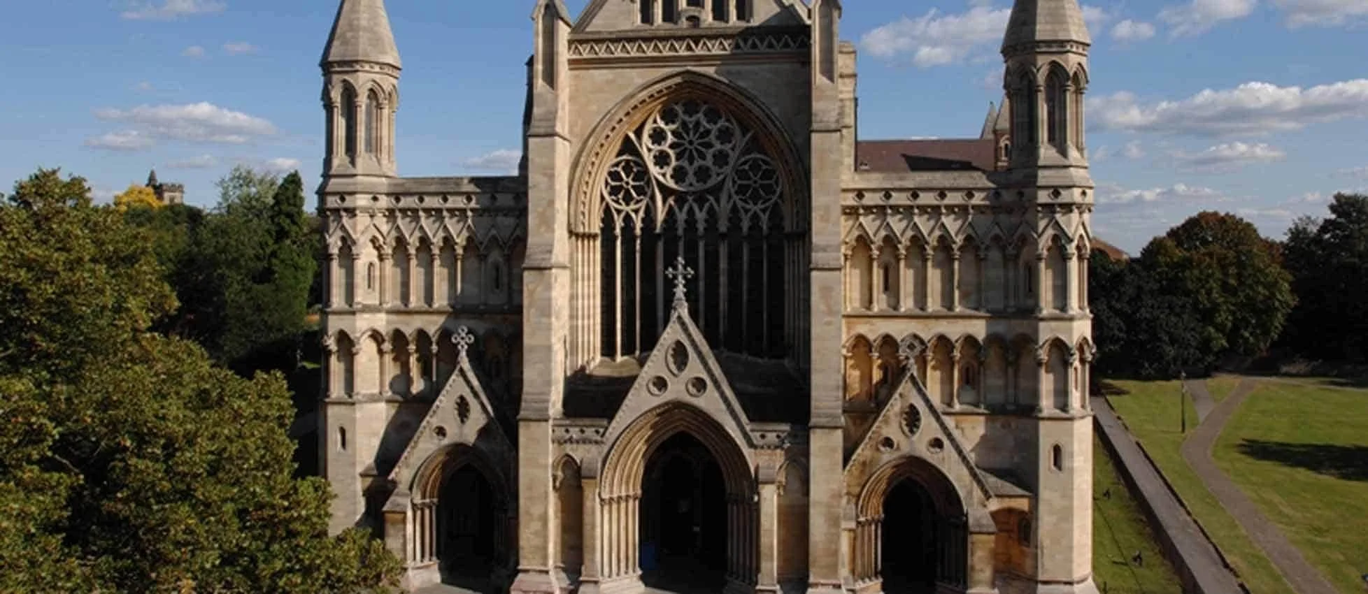 A historic Gothic-style church with pointed arches, large stained glass windows, ornamented stonework, and tall spires, surrounded by trees and a grassy area under a partly cloudy sky.