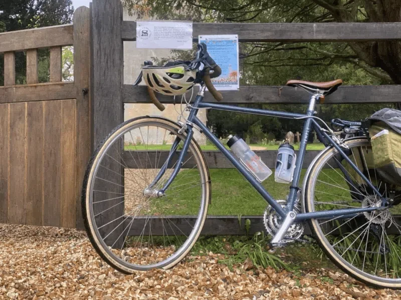 A blue bicycle parked against a wooden fence with a helmet hanging on the handlebars and a water bottle attached to the frame. The bicycle also has a rear bag and a small pouch, and is situated on a gravel pathway in a park with trees and grassy areas in the background.