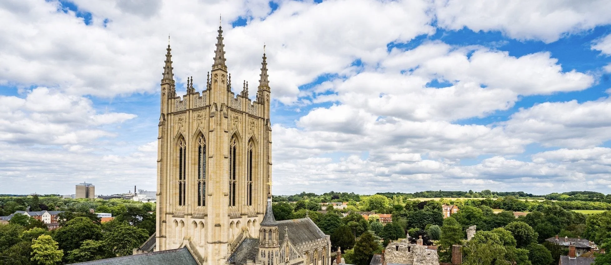 A tall Gothic-style church tower with pointed spires against a partly cloudy sky, overlooking a green cityscape with trees and buildings.