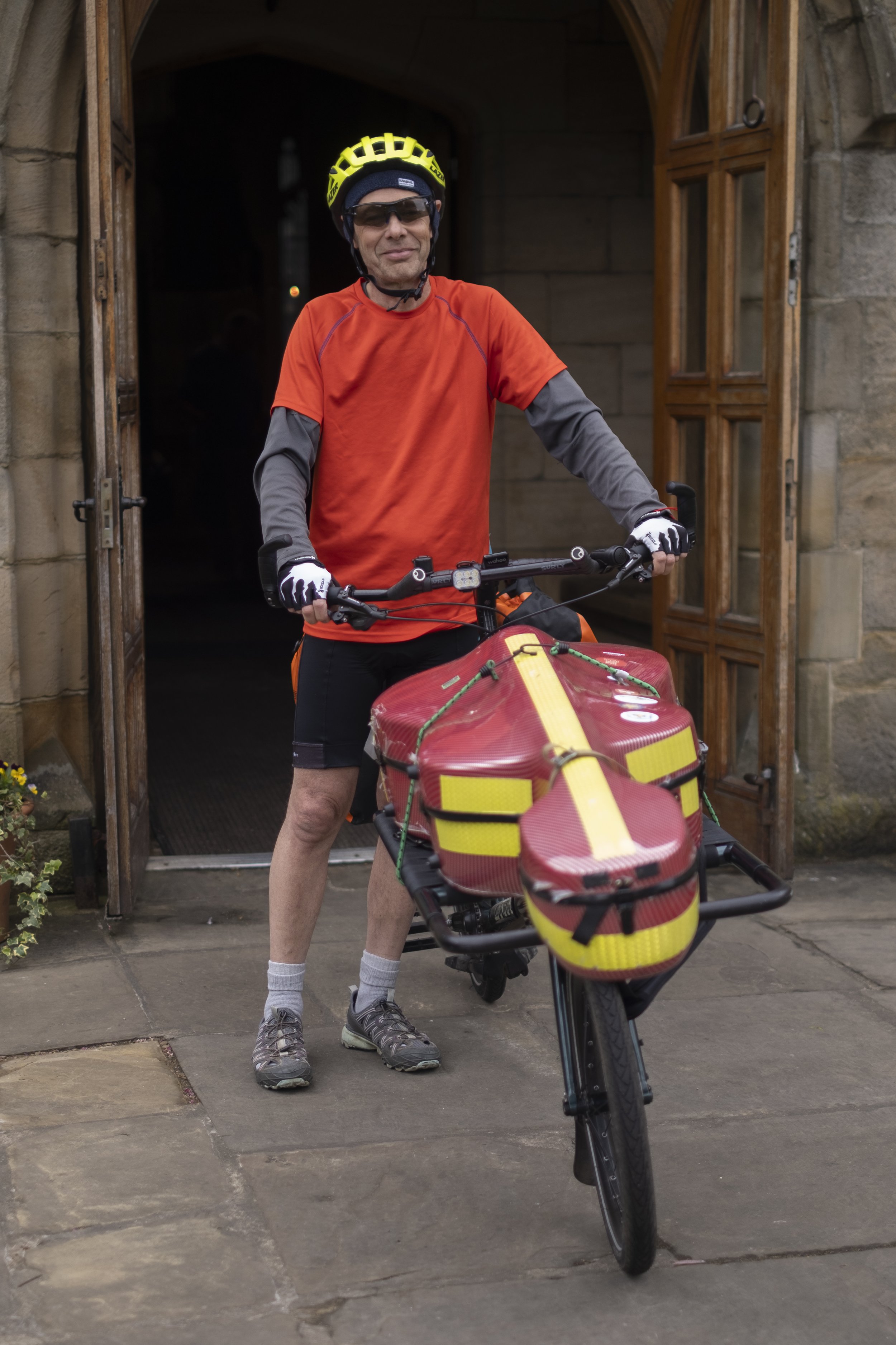 A man standing with a bike equipped with a red and yellow emergency medical bag in front of a stone building entrance.