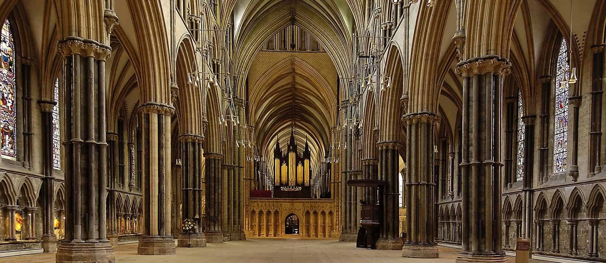 Interior view of a historic cathedral with tall pointed arches, stone columns, stained glass windows, and an organ at the altar.