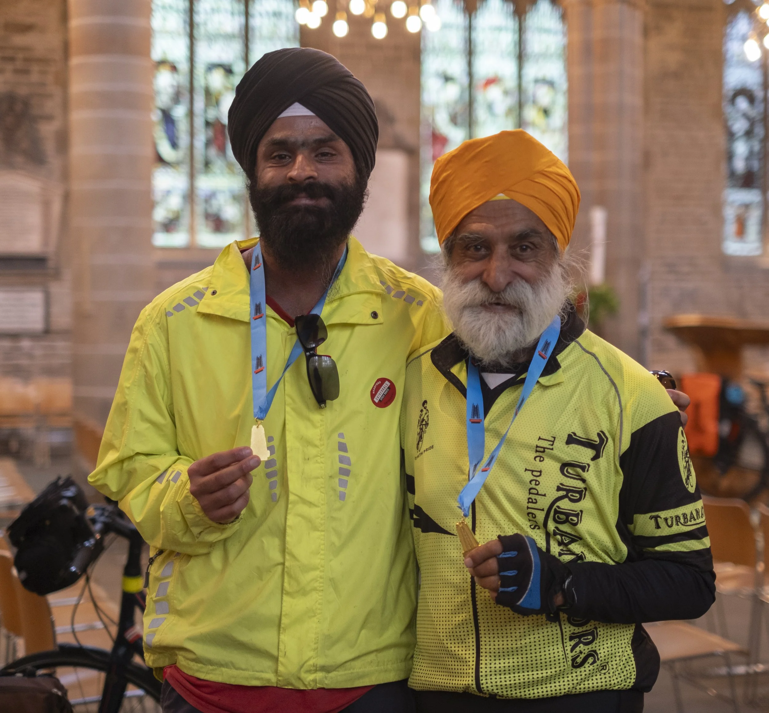 Two men wearing yellow cycling jackets with medals hanging around their necks inside a church or cathedral. One has a black turban, beard, and sunglasses hanging from his jacket. The other has a yellow turban and a white beard.