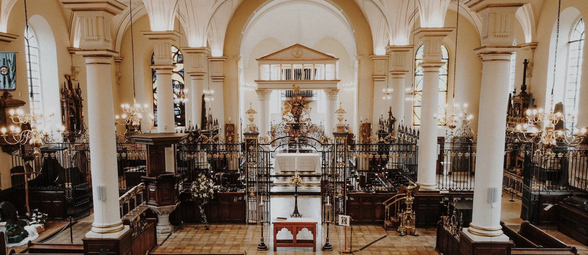 Interior of a church with tall white columns, stained glass windows, chandeliers, and ornate metalwork.