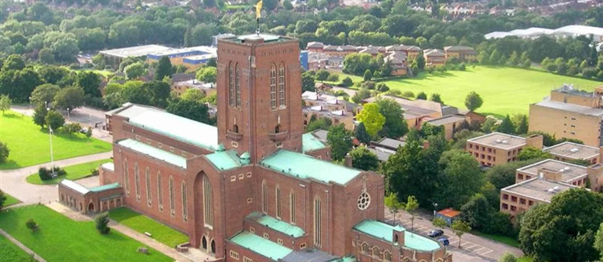 Aerial view of a large red brick church with tall windows and green metal roofs, surrounded by green lawns, trees, and nearby residential buildings in a small urban area.